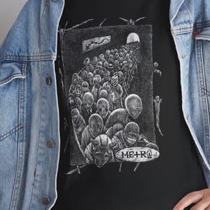 May include: Black t-shirt featuring a detailed black and white illustration of a crowd of figures with blank faces, some wearing masks. The word "METRO" is visible on a sign. The shirt is worn with a blue denim jacket.