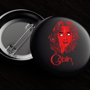 Puede incluir: Un botón negro con una ilustración roja de la cara de una mujer con un tercer ojo y la palabra "Goblin" en letras rojas que gotean.