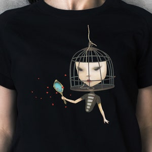 K&ouml;nnte beinhalten: Schwarzes T-Shirt mit einem Motiv einer Frau mit einem K&auml;fig &uuml;ber dem Kopf, die einen Spiegel h&auml;lt. Die Frau tr&auml;gt ein gestreiftes Shirt und hat rote Lippen. Der Hintergrund ist schwarz mit roten Herzen, die verstreut sind.