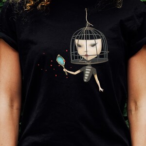 K&ouml;nnte beinhalten: Schwarzes T-Shirt mit einem grafischen Druck einer Frau mit einem Vogelk&auml;fig auf dem Kopf. Sie h&auml;lt einen Spiegel und rote Herzen schweben um sie herum.