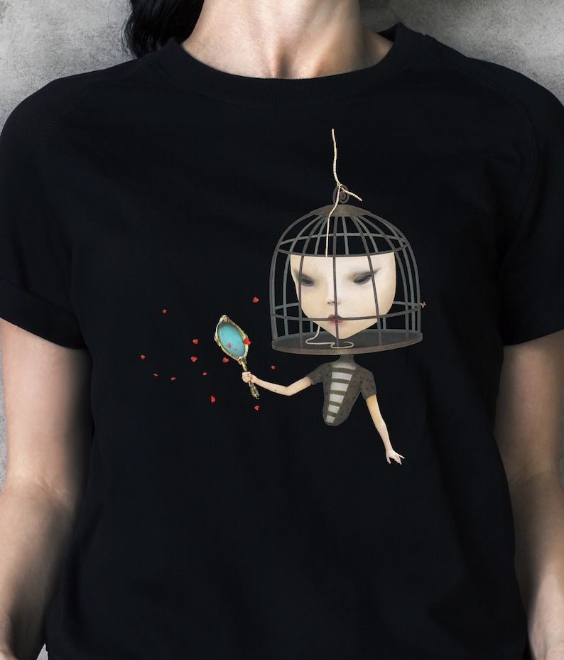 K&ouml;nnte beinhalten: Schwarzes T-Shirt mit einem Motiv einer Frau mit einem K&auml;fig &uuml;ber dem Kopf, die einen Spiegel h&auml;lt. Die Frau tr&auml;gt ein gestreiftes Shirt und hat rote Lippen. Der Hintergrund ist schwarz mit roten Herzen, die verstreut sind.