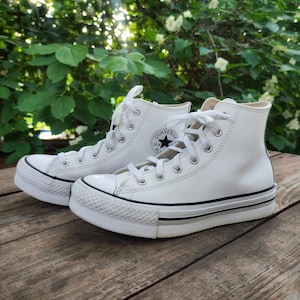 May include: A pair of white high-top platform sneakers with white laces. The sneakers have a black stripe along the sole and a black Converse All Star logo on the side. The shoes are on a wooden surface.
