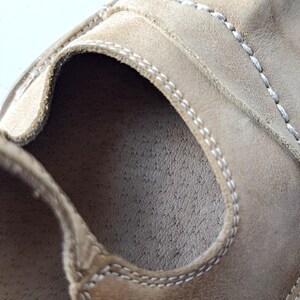 May include: Close-up of a tan leather shoe interior. The shoe features visible stitching in a contrasting white thread along the edges and seams. The interior has a textured surface, suggesting a focus on comfort and durability.