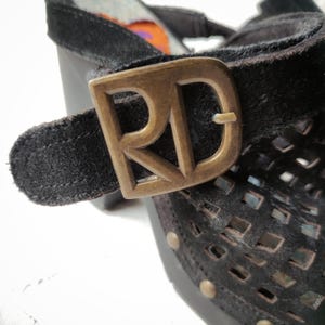 May include: Close-up of a black suede sandal with a gold-toned buckle featuring the letters "RD". The sandal has a woven pattern and is adorned with small metal studs.