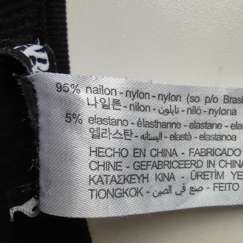 Puede incluir: Primer plano de una etiqueta de tela blanca con texto negro. La etiqueta indica una composici&oacute;n de material de 95% nailon y 5% elastano. Informaci&oacute;n adicional de fabricaci&oacute;n en varios idiomas, incluyendo espa&ntilde;ol y chino.