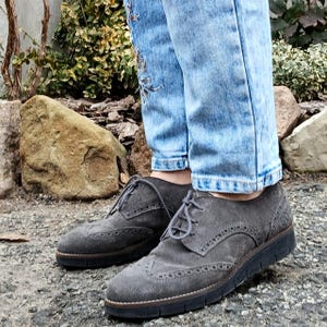 May include: Gray suede brogue shoes with a lace-up closure and a wingtip design. The shoes have a black rubber sole and are worn with light blue jeans.
