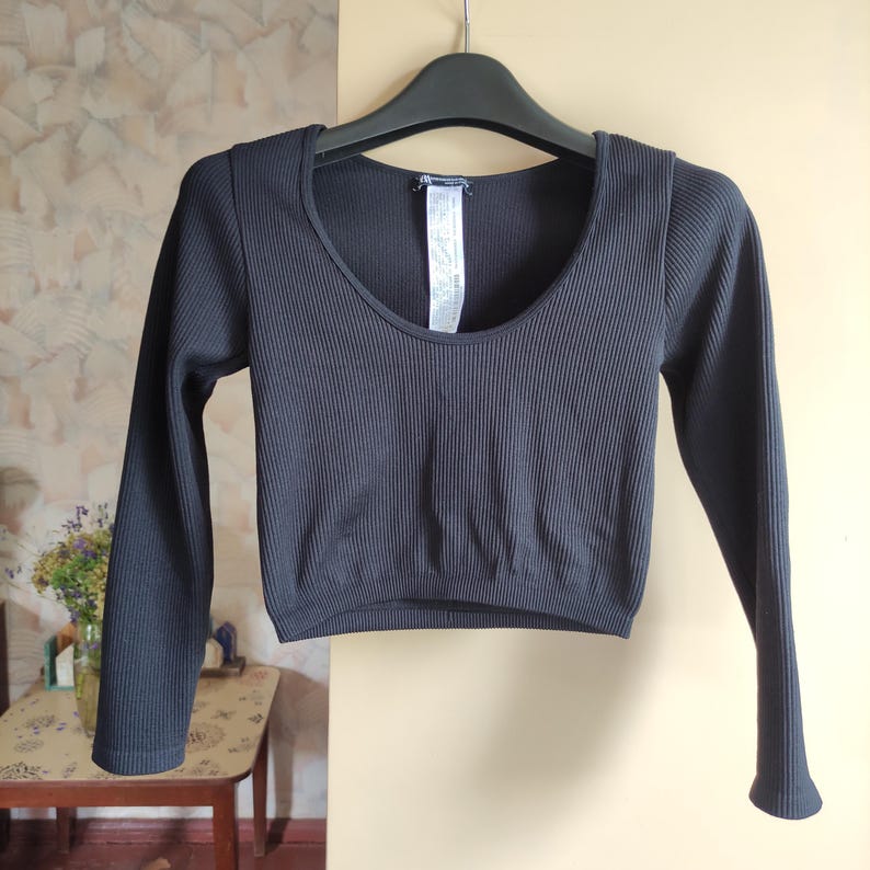 Puede incluir: Top corto negro acanalado con mangas largas y cuello redondo. La parte superior est&aacute; colgada de una percha negra. La tela tiene acanalado vertical. La parte superior es de color s&oacute;lido y parece estar hecha de un material el&aacute;stico.