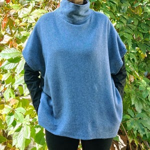 May include: A pale blue, short-sleeved sweater with a high roll neck. The sweater has a relaxed fit. The sleeves are short, and the sweater is worn over a black long-sleeved top. The background features green and red foliage.