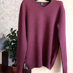 May include: A burgundy V-neck sweater. The sweater has long sleeves and a ribbed hem and cuffs. The neckline is a deep V-shape. The sweater is made of a soft, textured material. The sweater is a solid colour.