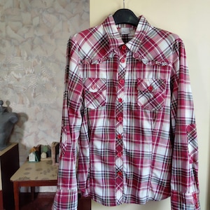 May include: A long-sleeved plaid shirt in shades of red, white, and gray, with two chest pockets and red buttons. The shirt has a collared neckline and decorative ruffles along the yoke. The sleeves have buttoned cuffs.