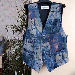 May include: A patterned vest in shades of blue, gray, and brown, featuring an abstract design. The vest has a V-neck, button closure, and two front pockets. The label "Angelo Lino" is visible on the inner lining.