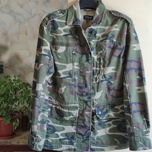 May include: A camouflage print jacket with a button-up front, two chest pockets, and two lower pockets. The jacket features a collar and is made of a green, brown, and black camouflage pattern. The jacket is hanging on a hanger.