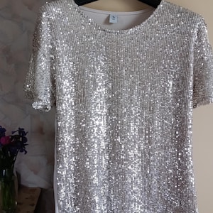 May include: A short-sleeved shirt covered in shimmering silver sequins on the front panel. The garment features a round neckline and is displayed on a black plastic hanger. The back and side panels are made of a plain, solid white fabric material.