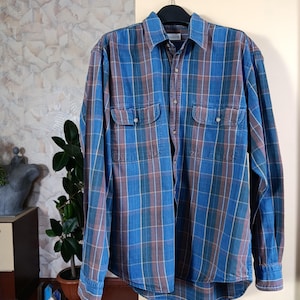 May include: A blue, brown, and green plaid button-down shirt with two chest pockets. The long-sleeved shirt is hanging on a black hanger. The shirt has a classic collar and buttoned cuffs.