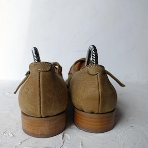 May include: A pair of tan leather Mary Jane shoes with a low wooden heel. The shoes have a strap across the top and are shown from the back, with shoe trees inserted. The leather has a textured appearance.