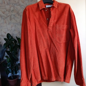 May include: An orange corduroy long-sleeve shirt with a collared neckline and a single chest pocket. The shirt has a button-down placket and a relaxed fit. The fabric has a ribbed texture.