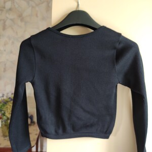 Puede incluir: Un top negro de punto acanalado de manga larga colgado de una percha negra. El top tiene un escote redondo y una longitud recortada. La tela tiene canal&eacute; vertical y las mangas son ajustadas.