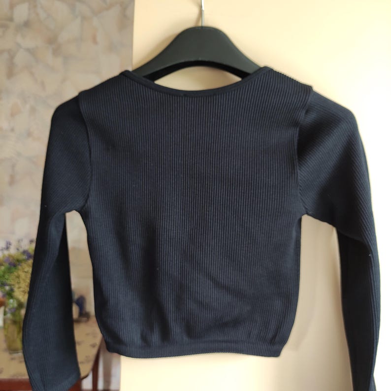 Puede incluir: Un top negro de punto acanalado de manga larga colgado de una percha negra. El top tiene un escote redondo y una longitud recortada. La tela tiene canal&eacute; vertical y las mangas son ajustadas.
