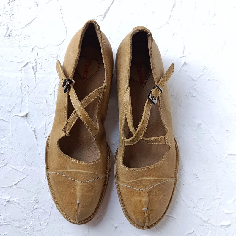 May include: A pair of tan leather Mary Jane style shoes. The shoes have a strap across the top with a buckle closure and a crisscross strap design. The shoes have a rounded toe and decorative stitching.