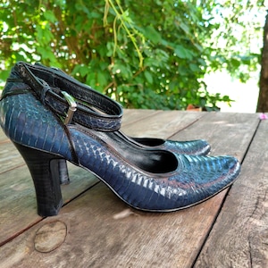 May include: A pair of blue high-heeled shoes with a snakeskin pattern. The shoes have a strap around the ankle and a black heel. The shoes are on a wooden surface.