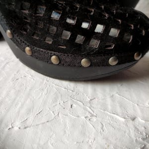 May include: Black leather clog with a cut-out design and metal accents. The clog has a thick sole and a rounded toe.