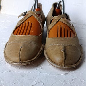 May include: A pair of tan leather Mary Jane style shoes with a strap and buckle closure. The shoes have a unique split-toe design and are displayed with wooden shoe trees. The shoes are on a white textured surface.
