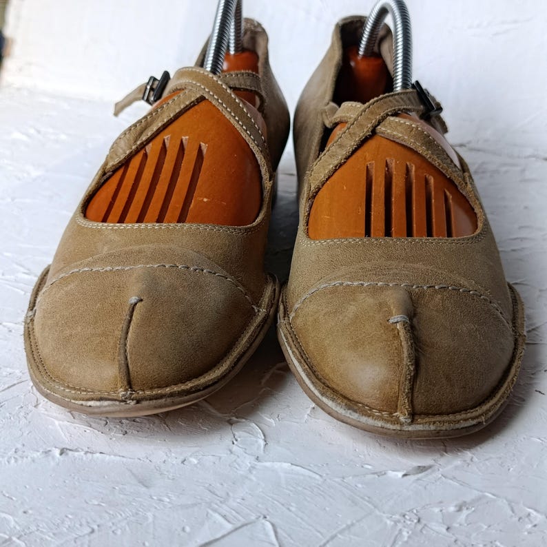 May include: A pair of tan leather Mary Jane style shoes with a strap and buckle closure. The shoes have a unique split-toe design and are displayed with wooden shoe trees. The shoes are on a white textured surface.