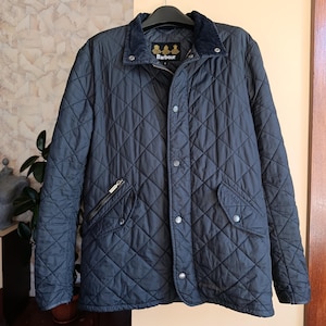 May include: A navy blue quilted jacket with a corduroy collar and a diamond pattern. The jacket features a zip and button closure, two front pockets, and the Barbour logo on the collar.