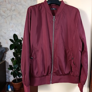 May include: A burgundy bomber jacket with a silver zipper and two front pockets. The jacket has a ribbed collar, cuffs, and hem. The jacket is hanging on a black hanger.