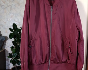 May include: A burgundy bomber jacket with a silver zip and two front pockets. The jacket has a ribbed collar, cuffs, and hem. The jacket is hanging on a black hanger.