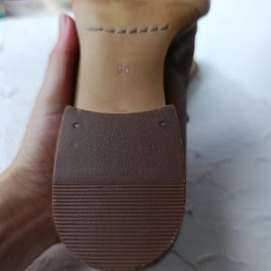 May include: Close-up of the sole of a brown shoe. The sole is made of a textured, dark brown rubber material. The heel area has the number "36" printed on it. The shoe is being held by a person.