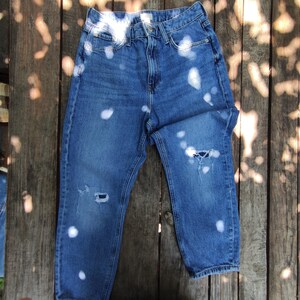 May include: Blue denim jeans with white bleach spots and distressed details. The jeans have a classic five-pocket design and a button and zipper closure. The jeans are laid flat on a wooden surface.