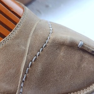 May include: Close-up of a tan leather slipper with white stitching. The slipper has a textured surface and a rounded toe. The stitching is visible along the seams and around the edges of the slipper. The slipper is on a wooden shoe form.