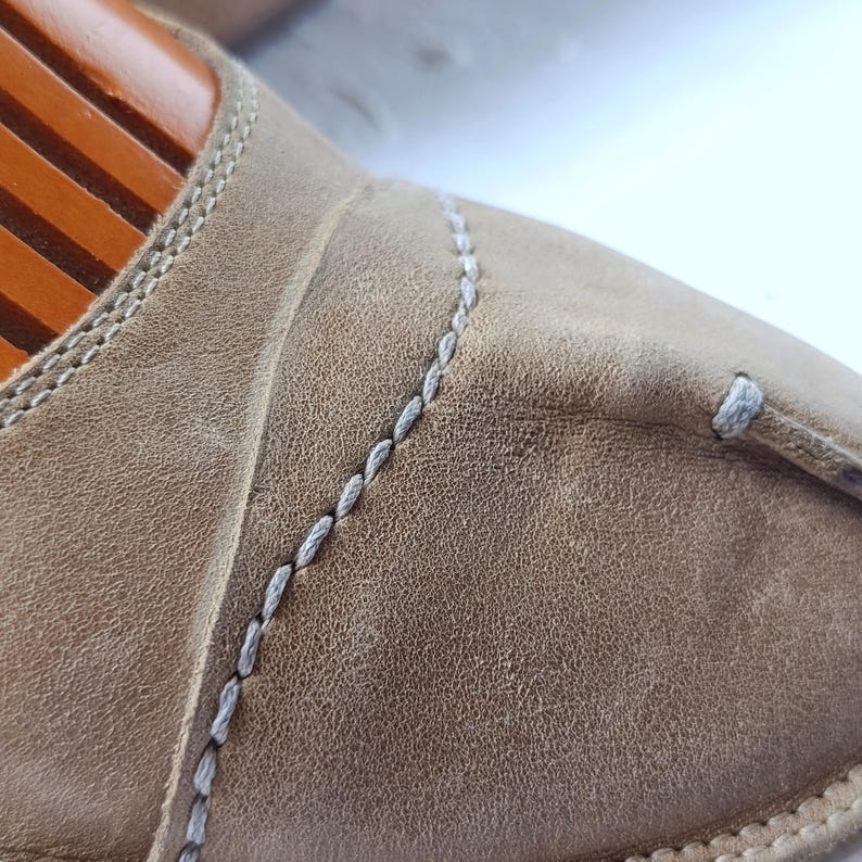 May include: Close-up of a tan leather slipper with white stitching. The slipper has a textured surface and a rounded toe. The stitching is visible along the seams and around the edges of the slipper. The slipper is on a wooden shoe form.