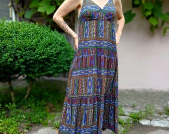 May include: A long, tiered maxi dress with a V-neck and spaghetti straps. The dress features a vibrant, repeating pattern in shades of blue, red, green, and gold. The dress is worn with brown sandals.