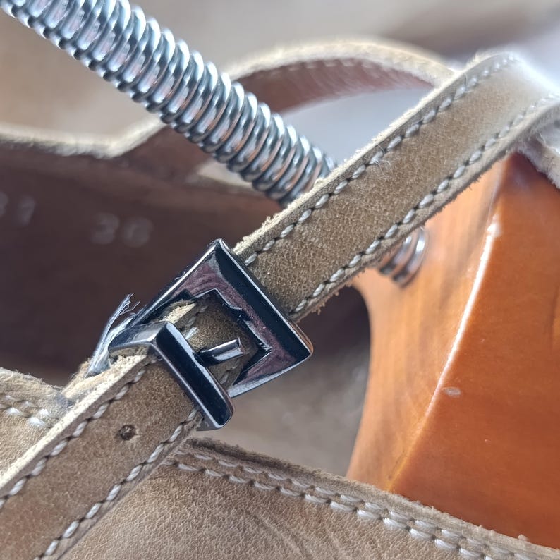 May include: Close-up of a tan leather sandal with a silver buckle. The sandal features a wooden sole and a strap with a silver buckle. The stitching is visible on the leather strap. The sandal is a size 36.