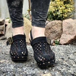 May include: Black suede clogs with a cut-out design and a buckle closure. The clogs have a wooden heel and a platform sole.