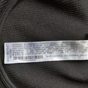 Puede incluir: Primer plano de una tela acanalada verde oscuro con una etiqueta blanca. La etiqueta incluye texto en varios idiomas, un c&oacute;digo de barras y la marca "INDITEX&reg;". La tela tiene una apariencia texturizada.