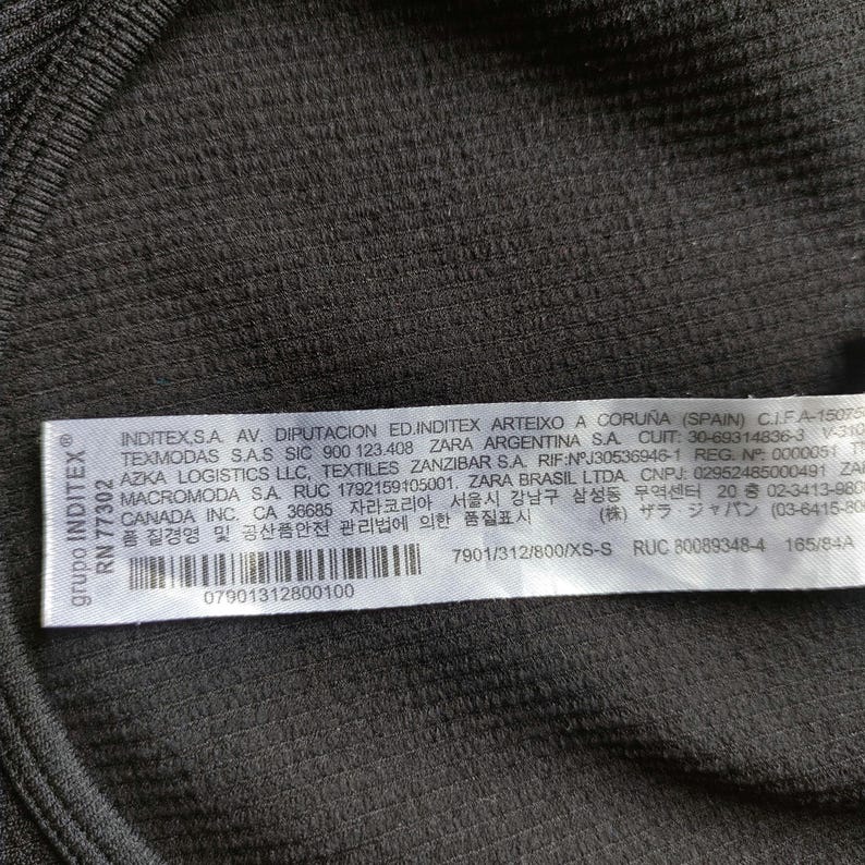 Puede incluir: Primer plano de una tela acanalada verde oscuro con una etiqueta blanca. La etiqueta incluye texto en varios idiomas, un c&oacute;digo de barras y la marca "INDITEX&reg;". La tela tiene una apariencia texturizada.