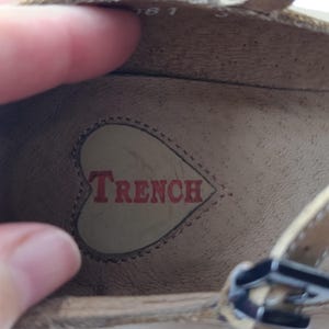 May include: Close-up of the inside of a beige shoe. The word "TRENCH" is printed in red inside a heart-shaped design. The shoe has a strap with a silver buckle.