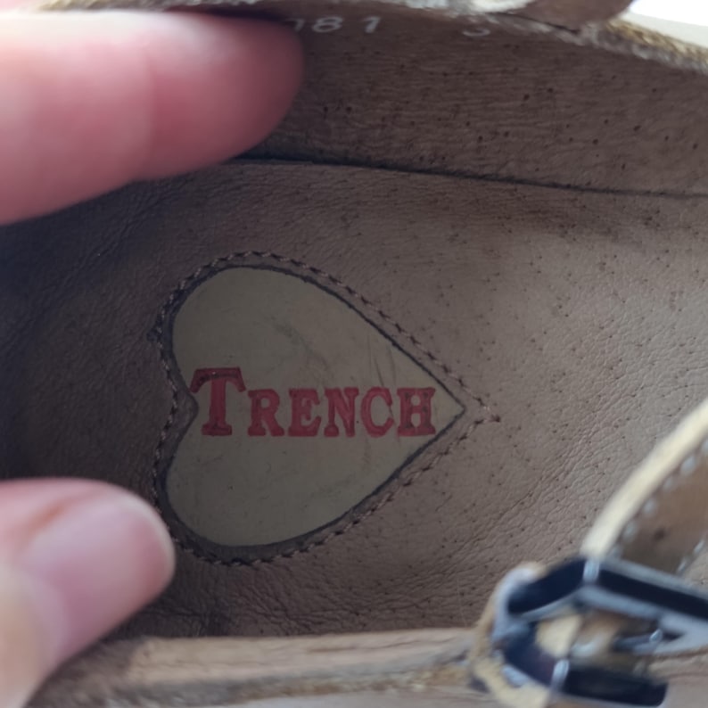 May include: Close-up of the inside of a beige shoe. The word "TRENCH" is printed in red inside a heart-shaped design. The shoe has a strap with a silver buckle.