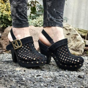 May include: Black suede heeled clogs with a cut-out design and a buckle with the initials "RD".
