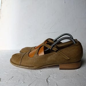 May include: A pair of olive green Mary Jane style shoes with a strap and buckle closure. The shoes have a rounded toe and a low, stacked heel. Shoe trees are inserted in the shoes.
