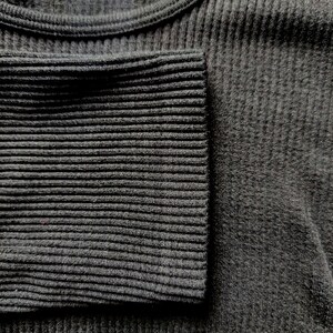 Puede incluir: Primer plano de una camiseta de manga larga de punto acanalado gris oscuro. El pu&ntilde;o de la manga es visible, mostrando un patr&oacute;n de canal&eacute; m&aacute;s apretado. La tela parece suave y c&oacute;moda.