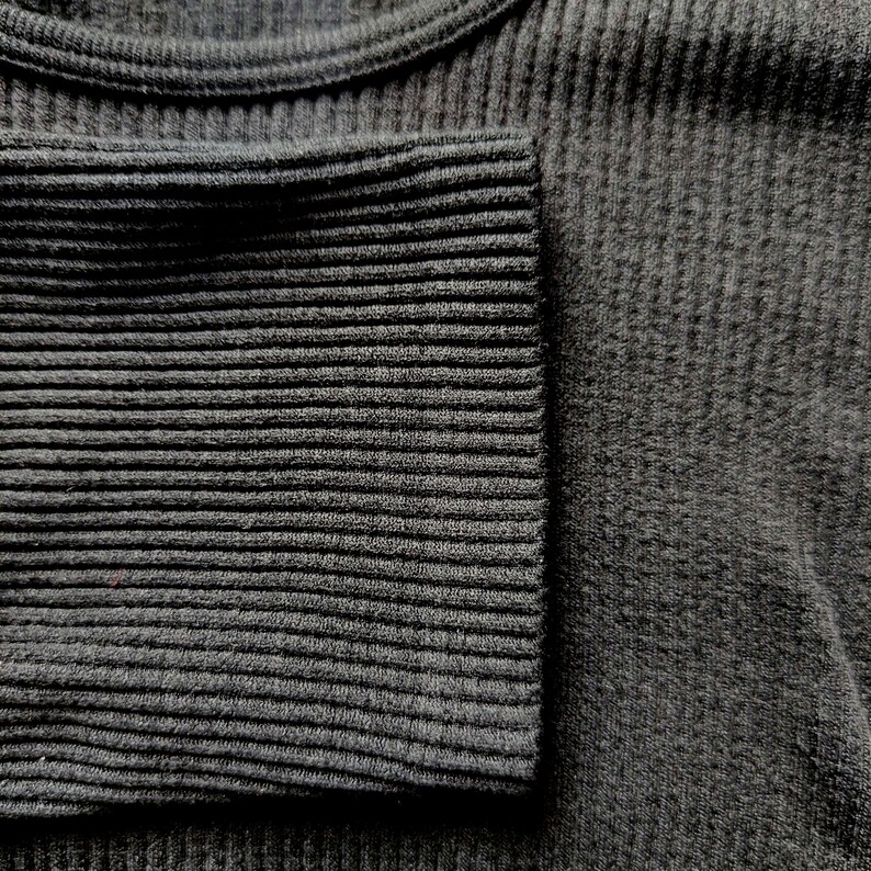 Puede incluir: Primer plano de una camiseta de manga larga de punto acanalado gris oscuro. El pu&ntilde;o de la manga es visible, mostrando un patr&oacute;n de canal&eacute; m&aacute;s apretado. La tela parece suave y c&oacute;moda.