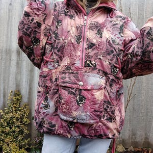 May include: A pink and burgundy anorak jacket with a floral and abstract pattern. The jacket features a front pocket, a zipper closure, and a hood. The anorak has a drawstring at the bottom and around the hood. The text "MASSAGE" is visible on the front.