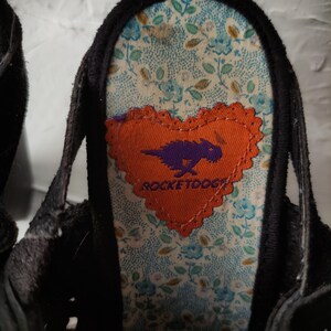 May include: Close-up of the inside of a black shoe with a floral pattern. A heart-shaped orange patch with a purple cartoon creature and the text "ROCKETDOGG" is sewn onto the shoe.