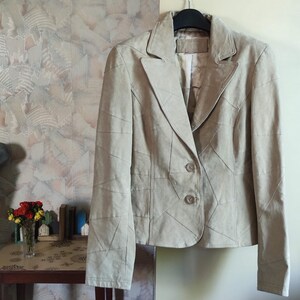 May include: A beige suede blazer with a patchwork design, hanging on a black hanger. The jacket has a notched collar, two buttons, and a tailored fit. The background features a patterned wall and a small table with flowers.