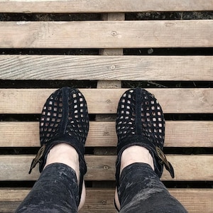 May include: A pair of black leather sandals with a grid pattern and buckle closures. The sandals are worn by a person with black pants.