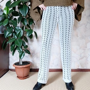 May include: White, open-knit pants with a flared leg. The pants have a detailed, patterned design. The person is wearing black shoes and a brown sweater. A potted plant is in the background.